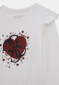 T-shirt in cotone organico bianco con maniche corte arricciate, caratterizzato da un design a forma di cuore in glitter rosso e lettere nere circondate da piccole stelle rosse.