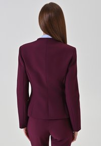 Bordeaux op maat gemaakt blazer met een slanke silhouet, zachte stof en gestructureerde schouders; voorzien van een blauwe kraag die eronder vandaan piekt.