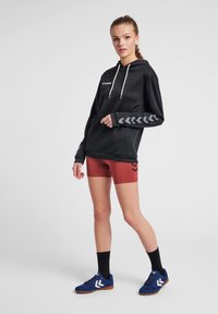 Felpa nera con cordini bianchi e maniche grigie a motivo, abbinata a pantaloni corti color ruggine e scarpe sportive blu con dettagli bianchi.