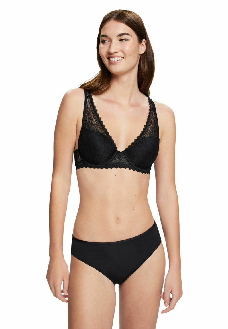 Schwarzer Spitzen-BH mit tiefem V-Ausschnitt, welligen Kanten und strukturiertem Muster. Kombiniert mit schlichten schwarzen Bikini-Unterteilen.