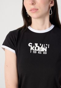Black cotton t-shirt with white trim and Calvin Klein 1968 logo in bold white letters. Close-up view, featuring short sleeves and smooth texture.