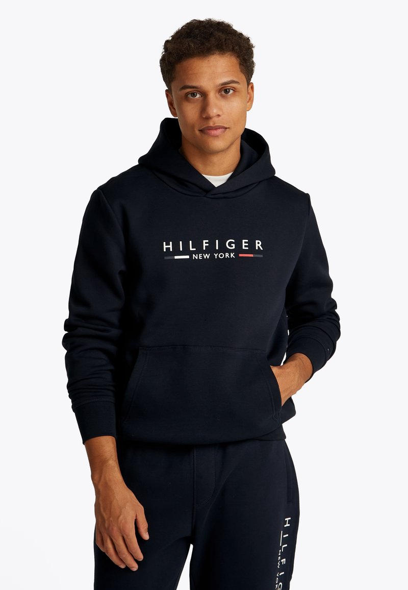 Tumši zils kapuci džemperis kokvilnas maisījumā ar priekšējā kabatu, uz kura ir uzraksts "HILFIGER NEW YORK" baltā un sarkanā krāsā uz krūtīm.