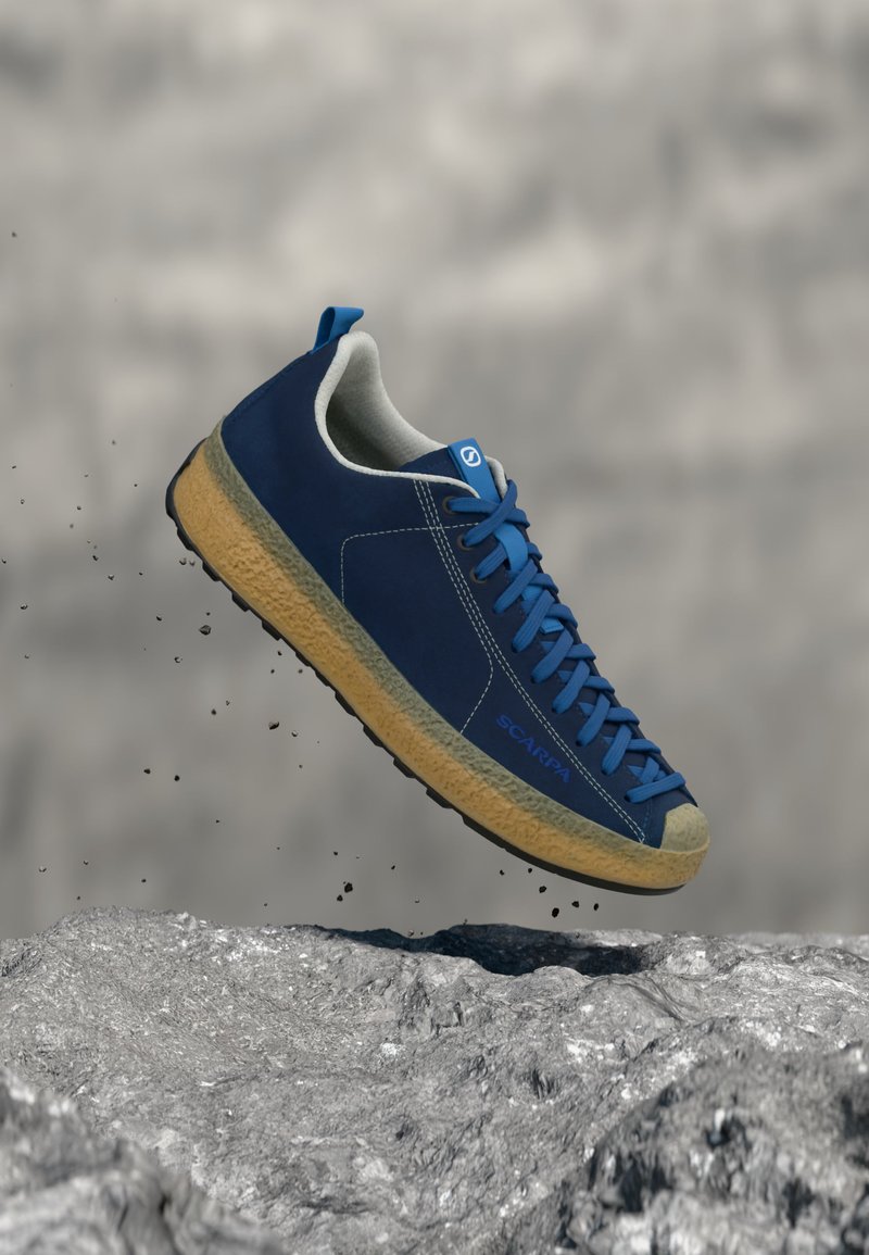 Μπλε αθλητικό παπούτσι με ανοιχτό καφέ σόλα, αιωρούμενο πάνω από βραχώδη επιφάνεια, που δείχνει λεπτομερή ραφή και λογότυπο στη πλευρά.