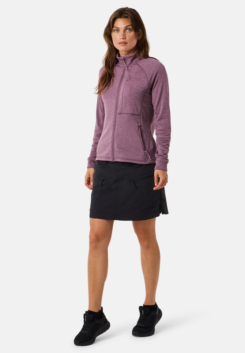 Frau steht in einer lila Reißverschlussjacke, einem schwarzen knielangen Rock mit Taschen und schwarzen Sportschuhen vor einem einfarbigen Hintergrund.