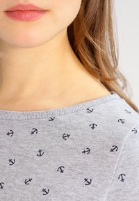 Textil gris con pequeños estampados de anclas azul marino. La camisa tiene un escote redondo y ribete acanalado a lo largo del cuello.