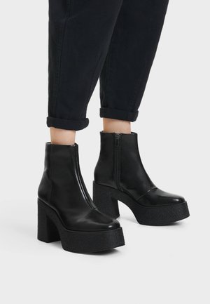 Personne portant des bottines noires à plateforme avec des talons carrés texturés et des fermetures éclair latérales, associées à un pantalon noir retroussé.