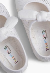 White textile shoes featuring a perforated design and a bow accent on the front. Includes a label reading "Pisamonas, Hecho en España."