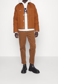 Giacca color ruggine con cuciture bianche e tasca frontale, indossata sopra un hoodie bianco. Pantaloni in corduroy marrone e stivali neri alla caviglia.