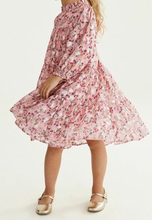 Jeune fille dans une robe rose à fleurs à manches longues en train de tournoyer, portant des chaussures Mary Jane dorées sur un fond uni.