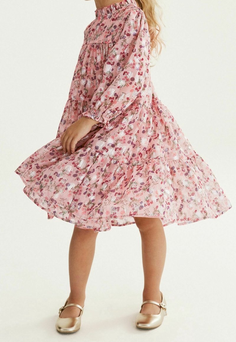 Jeune fille dans une robe rose à fleurs à manches longues en train de tournoyer, portant des chaussures Mary Jane dorées sur un fond uni.