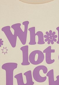 Krémová mikina s výrazným fialovým nápisom "Veľa šťastia" s kvetinovými a hviezdičkovými detailmi. Mäkká, textúrovaná látka.