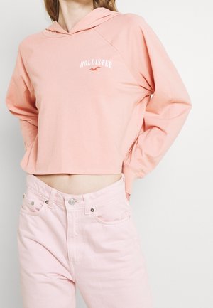 Kobieta w jasnoróżowej krótkiej bluzie z kapturem z logo "Hollister" i jasnoróżowych dżinsach z wysokim stanem, z rękami za plecami, na białym tle.