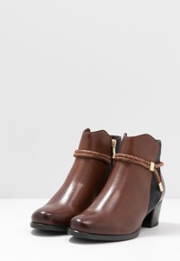 Bottes basses en cuir marron à finition brillante, avec un détail tressé et une fermeture éclair latérale, complétées par un panneau en textile bleu marine et des accents dorés.