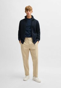 Giacca zip navy, pantaloni beige e sneakers bianche. La giacca ha un colletto alto, mentre i pantaloni presentano un solo bottone e una leggera svasatura.