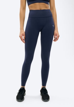 Weiblicher Oberkörper und Beine in marineblauen Leggings mit hohem Bund und schwarzen Sportschuhen, stehend vor einem schlichten weißen Hintergrund.