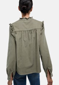 Chemise vert olive avec des accents d'épaules froncés, manches longues et coupe ample. Tissu texturé avec une apparence légèrement froissée.