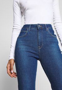 Högmidjade blå denimjeans med en slät textur, fem fickor och kontrasterande sömmar, med knappstängning fram och logotypdetalj.