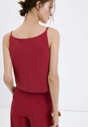 Vrouw met vastgebonden haar die een mouwloos rood topje en bijpassende broek draagt, geaccentueerd met meerdere gouden armbanden aan de rechterpols.