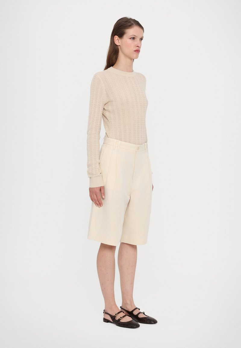 Beżowa, prążkowana bluzka z długim rękawem w połączeniu z kremowymi, krótkimi spodniami do kolan z guzikami i falbankami. Model nosi ciemnobrązowe buty na obcasie z otwartym piętą.