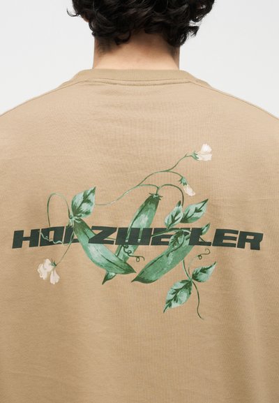 Πίσω όψη ατόμου που φοράει μπεζ πουκάμισο με πράσινους λοβούς αρακά, φύλλα, λευκά λουλούδια και σκούρο πράσινο κείμενο "HILZWEILER" στην πλάτη.