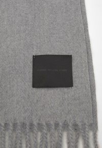 Siva volnena šal z mehkimi vlakni, z resicami na koncih. Na enem robu je pritrjen črn etiket z vtisnjenim besedilom.