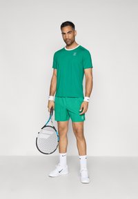 Πράσινο αθλητικό μπλουζάκι με λευκό γιακά, σε συνδυασμό με ταιριαστά πράσινα σορτς. Αξεσουάρ περιλαμβάνουν βραχιολάκια και μια ρακέτα τένις.