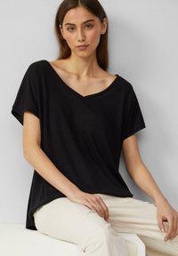 T-shirt noir à col en V avec des manches courtes, coupe décontractée, texture douce, porté avec un pantalon beige clair, sur fond neutre.