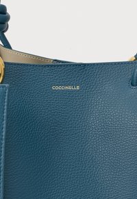 Kék texturált bőrtáska arany kiegészítőkkel, a "COCCINELLE" felirattal arany betűkkel a középső elülső részen.