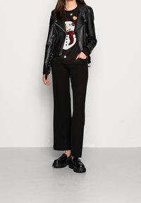 Černá kožená motocyklová bunda přes černý svetr s grafikou sněhuláka, doplněná o černé široké kalhoty a robustní černé polobotky.