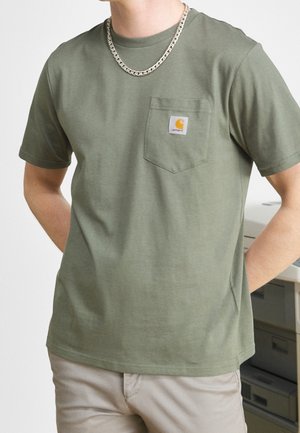 Person wearing olive green Carhartt pocket t-shirt and beige pants, with silver chain necklace, standing with hands behind back.