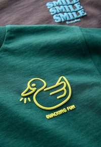 Sweatshirt en coton vert avec un graphique de canard brodé jaune et le texte "QUACKING FUN" en dessous. Texture lisse, design décontracté.