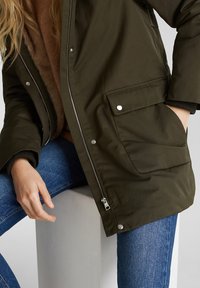 Femme portant une veste zippée vert foncé avec poches à boutons-pression, jean bleu et pull marron assise sur un cube blanc, une main dans la poche.