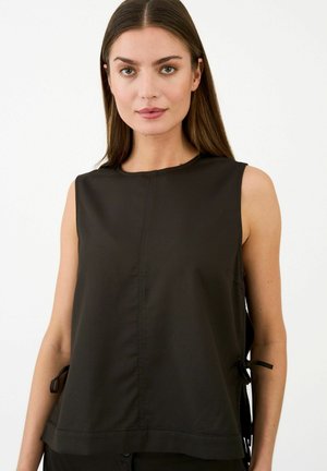 Femme aux cheveux bruns raides porte un haut noir sans manches avec des liens sur les côtés, debout devant un fond blanc uni.