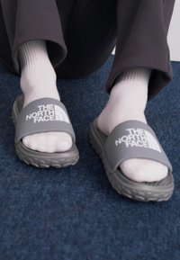 Grå slide-sandaler med en bred rem som har "THE NORTH FACE"-logotypen. Bärs över vita strumpor, placerade på en blå matta. Strukturerad gummisula.
