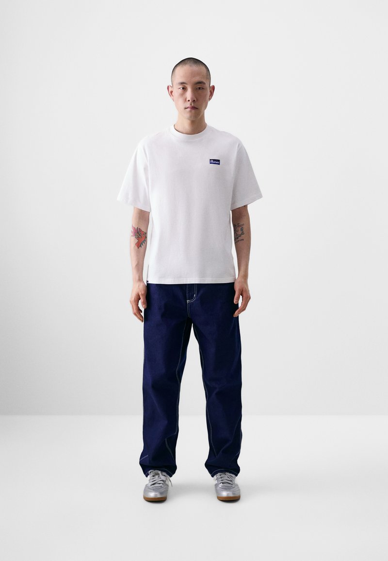 Camiseta branca de manga curta com um logotipo azul no peito esquerdo, combinada com calças de ganga azul marinho e ténis prateados. Design e cores simples.