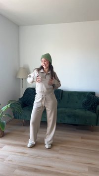 Conjunto de tela beige y suave que incluye un pulóver con cremallera y pantalones de chándal. Gorro de canalé verde. Suelo de madera clara y sofá de terciopelo verde de fondo.