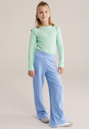 Camisa de manga larga verde claro con detalles en los hombros, combinada con pantalones anchos de pana azul con rayas verticales. Calzado con zapatos plateados.