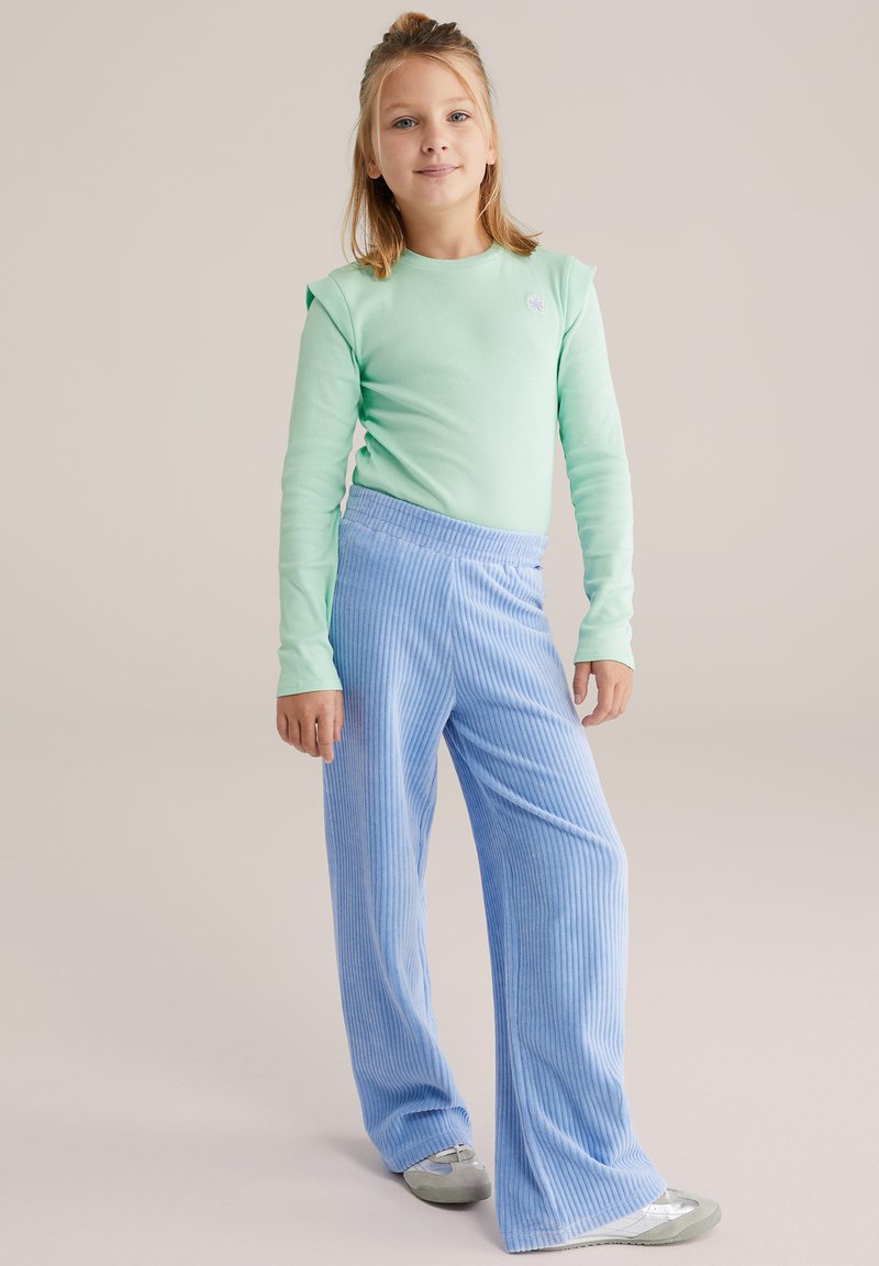 Világoszöld hosszú ujjú ing válldíszítéssel, kék kordbársony bő szárú nadrággal, amely függőleges csíkos mintázatú. Ezüst színű cipővel viselve.