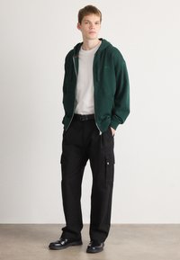 Grøn zip-up hoodie med en tekstureret overflade, båret over en hvid t-shirt. Parret med sorte cargo bukser med sidelommer og bælte. Sorte sko.