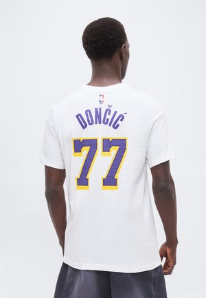 Homme portant un maillot NBA blanc avec le nom "Dončić" et le numéro "77" en violet et jaune dans le dos.