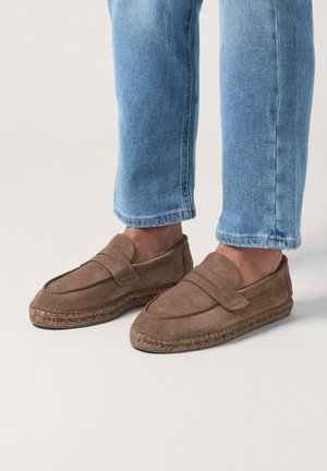 Braune Wildleder-Loafer mit geflochtenen Sohlen, getragen zu hochgekrempelten hellblauen Jeans vor einem schlichten Hintergrund.