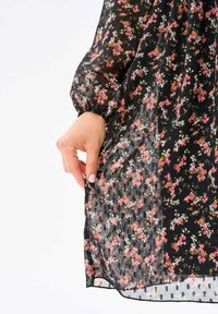 Käsi, joka pitää kiinni läpikuultavasta mustasta mekosta, jossa on vaaleanpunaisia ja valkoisia kukkakuoseja sekä elastiset hihansuut.