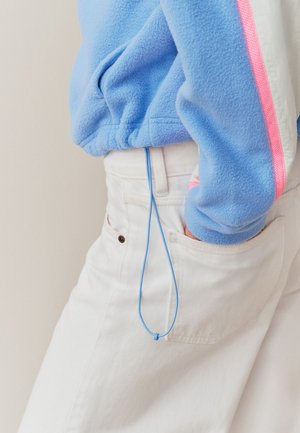Person, die weiße Hose trägt, mit einer Hand in der Tasche und einer blauen Fleecejacke mit pinkem Streifen, eine blaue Kordel, die vom Bund hängt.