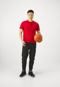 T-shirt vermelha de manga curta com logótipo preto, calças pretas justas e sapatilhas desportivas pretas/roxas. Segurando uma bola de basquete laranja.