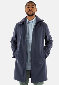 El abrigo impermeable de color navy presenta un cuello alto, botones a presión, bolsillos laterales y una textura suave, superpuesto a una camisa de cuadros en azul claro.