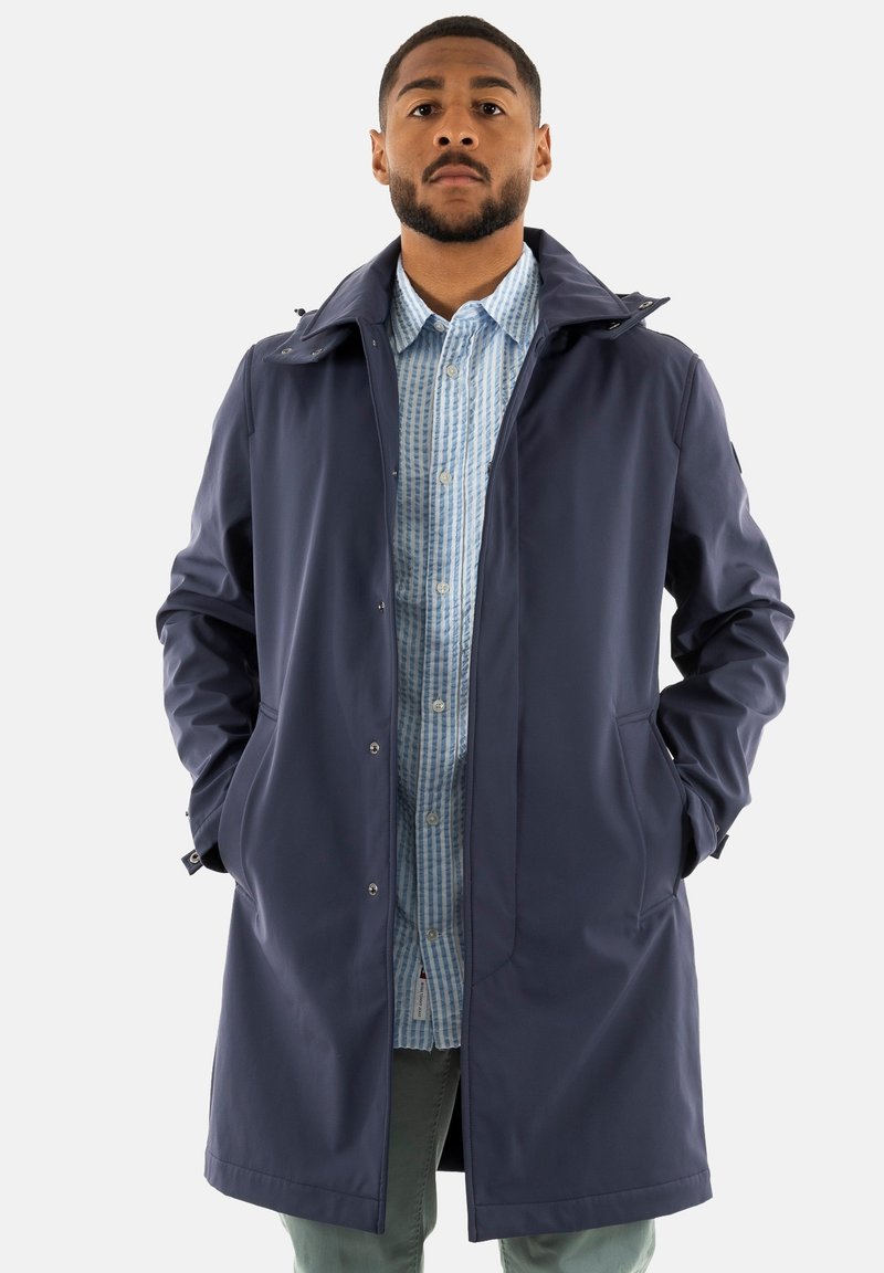 El abrigo impermeable de color navy presenta un cuello alto, botones a presión, bolsillos laterales y una textura suave, superpuesto a una camisa de cuadros en azul claro.