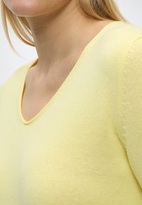 Gros plan sur une personne portant un pull en tricot jaune à col en V, montrant le col, l'épaule et la partie supérieure de la poitrine.