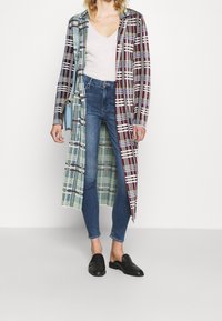 Abrigo largo de cuadros con patrones multicolores en rojo, azul, blanco y verde, llevado sobre jeans ajustados y zapatos negros sin cordones.