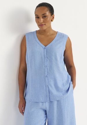 Kobieta o ciemnych włosach, ubrana w jasnoniebieską bezrękawnikową bluzkę z guzikami i dopasowane luźne spodnie, stoi przy białej ścianie.