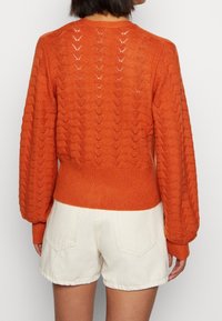 Pomarańczowy sweter dziewiarski z teksturowanym wzorem zygzaka, z prążkowanymi mankietami i dołem. W zestawieniu z jasnymi szortami.